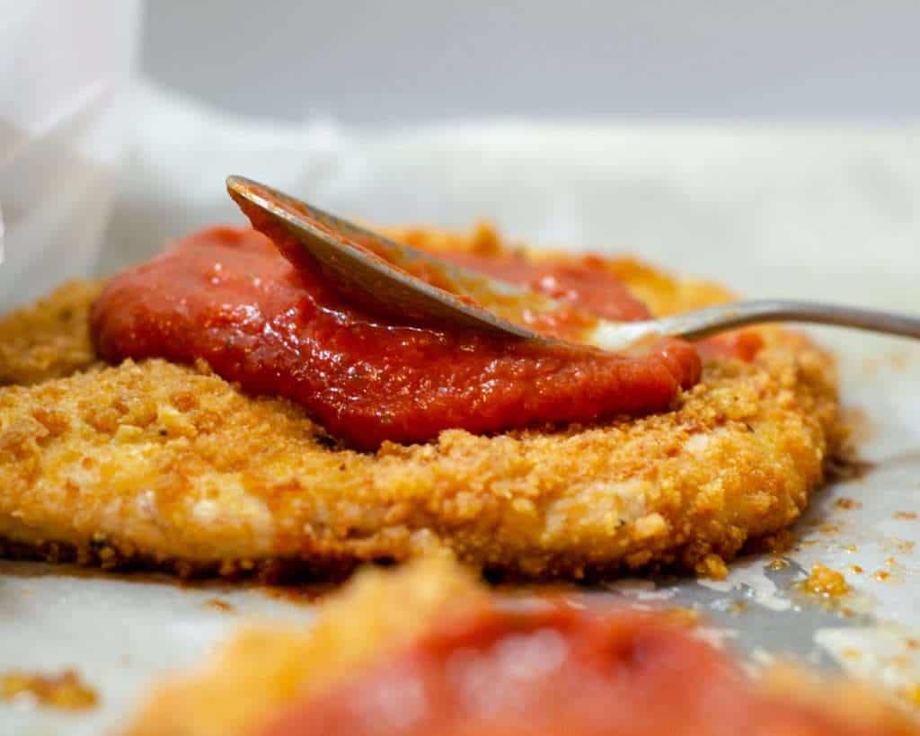 Crispy baked pork rind breaded chicken being topped with marinara