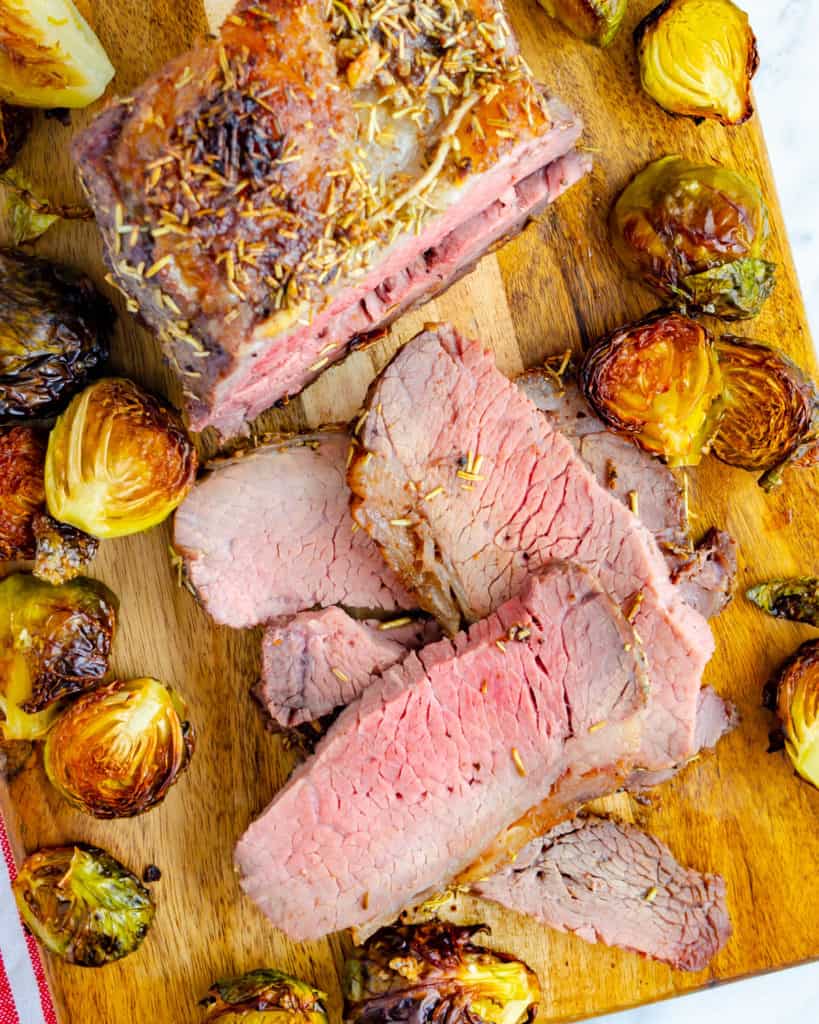 Slices of roast beef on a cutting board with roasted Brussels sprouts