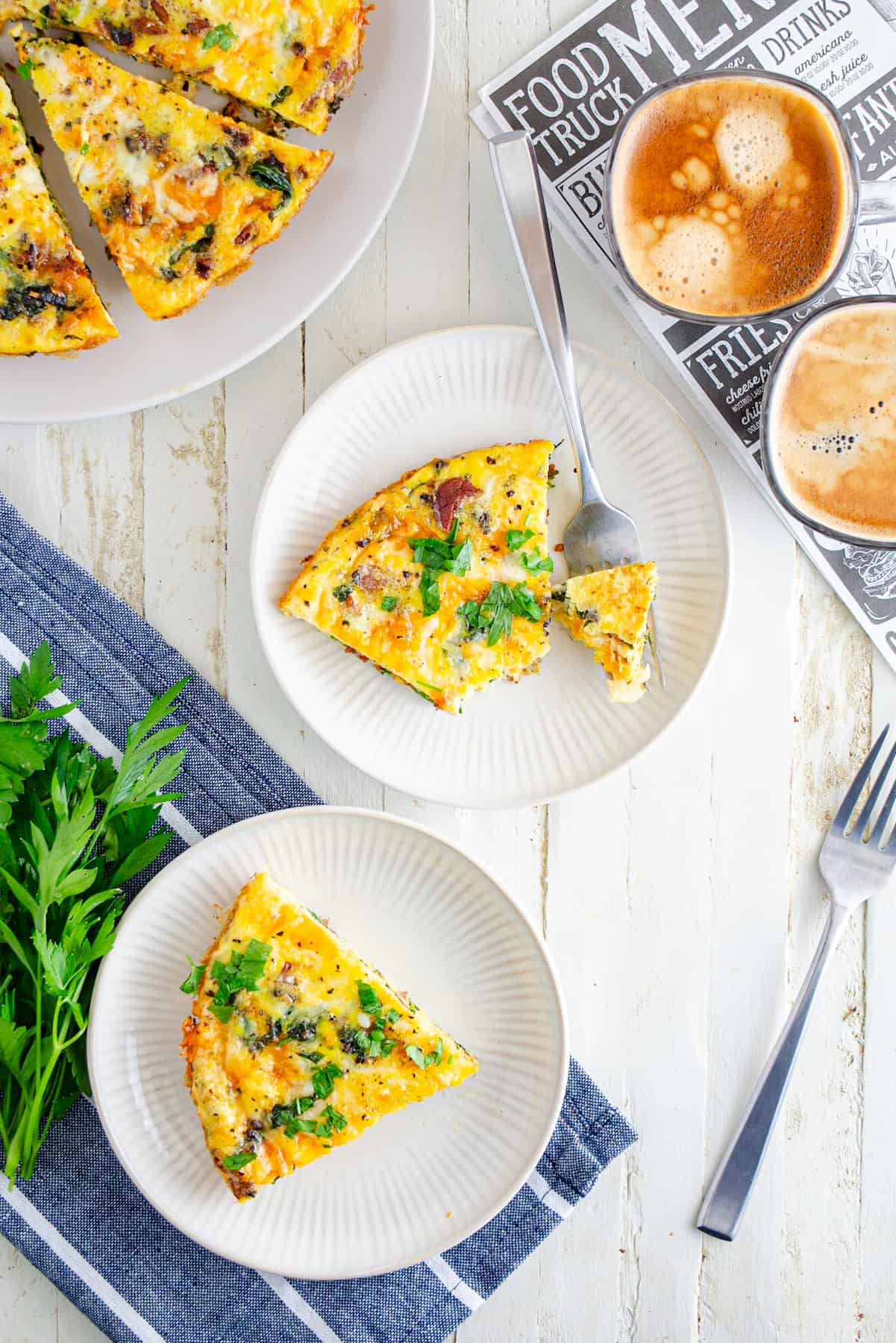 Slices of baked frittata on small white plates garnished with parsley and cheese with a side of lattes.