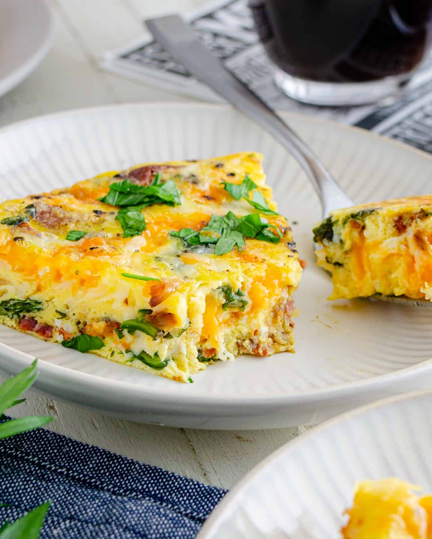 A slice of baked frittata on a plate with a bite out of it showing the beautiful cheese, spinach, and bacon inside.