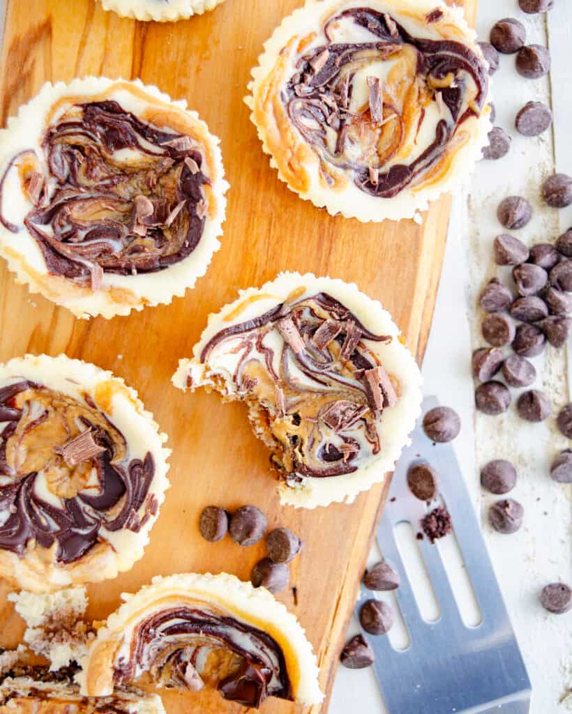 Mini muffin tin keto cheesecakes on a wooden board surrounded by low carb chocolate chips and peanut butter.