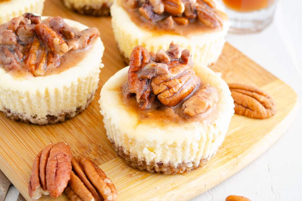 Keto maple pecan mini cheesecakes on a wooden cutting board garnished with fresh pecans.