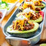 Chili cheese stuffed avocados fully loaded with cheese and sour cream on a metal tray surrounded by garnish with a grey napkin neatly folded beneath.